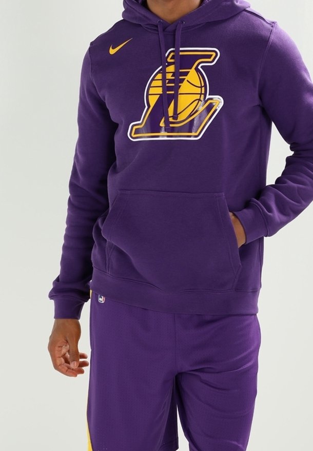 Personne portant un sweat à capuche violet et un short avec un logo de basketball jaune et une main dans la poche du sweat à capuche sur un fond uni.