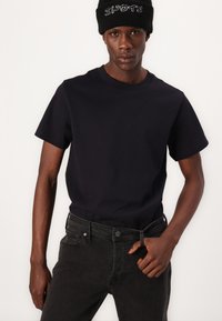 Camiseta de algodón azul marino con mangas cortas, combinada con pantalones vaqueros negros de mezclilla. El modelo lleva un gorro negro con detalles en texto blanco, posando frente a un fondo claro.