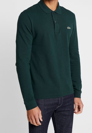 Homme portant un polo à manches longues vert foncé avec un petit logo de crocodile et un jean bleu foncé, sur un fond uni.