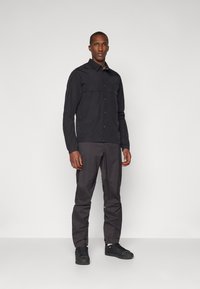 Black long-sleeve shirt with two chest pockets, paired with dark gray pants and black sneakers. Smooth texture, structured fit.