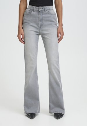 Kvinde iført lysegrå, højtaljede vide jeans med sort top og sorte spidse sko, stående mod en ensfarvet baggrund.