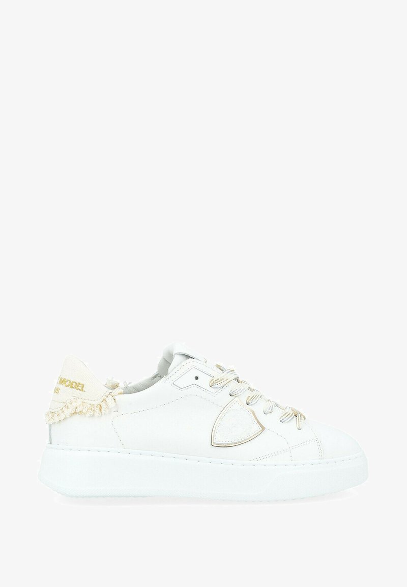 Sneaker bianchi in pelle con un accento in tessuto testurizzato, hardware dorato e frange decorative sul tallone. Suola in gomma spessa e dettagli cuciti.