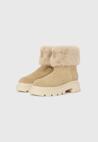 Bottines en suede beige avec un revers en fourrure et une semelle texturée. Présente un détail de logo et un design de semelle épaisse. Texture lisse sur l'ensemble.