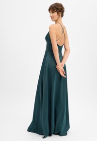 Femme en robe longue en satin vert sarcelle avec fines bretelles croisées dans le dos, debout les mains jointes derrière le dos.