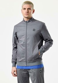 Grå zip-up jakke lavet af glat stof, med st�åkrave, sidelommer og ribbede manchetter. Sort logo-patch på brystet.