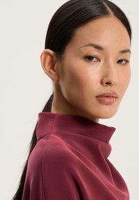 Top burgundy, cu guler înalt, realizat dintr-un material neted; are un croi lejer și detalii subtile pe umeri. Linii curate și design modern.
