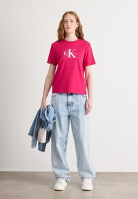Camiseta de algodón fucsia con un logo blanco, combinada con jeans de corte holgado en azul claro y zapatillas blancas. Una chaqueta de mezclilla está colgada sobre un brazo.
