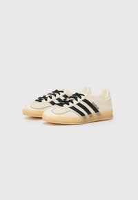 Beige sportskor med svarta ränder, strukturerad överdel och gummisula. Har snörning och "Gazelle"-logotyp på sidan.