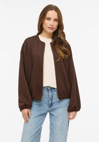 Pruun lukuga jope lõdva lõikega, soonikkoes varrukasäärtega ja tasase tekstuuriga, mida on kantud koos hele särk ja siniste teksapükstega.