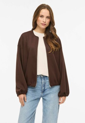 Brun zip-up jacka med avslappnad passform, ribbade muddar och slät textur, kombinerad med en ljus skjorta och blå jeans i denim.