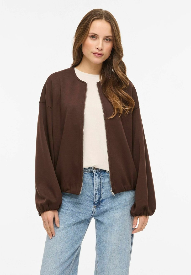 Pruun lukuga jope lõdva lõikega, soonikkoes varrukasäärtega ja tasase tekstuuriga, mida on kantud koos hele särk ja siniste teksapükstega.