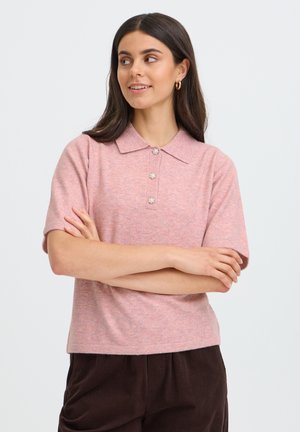 Giovane donna con lunghi capelli scuri che indossa una camicia rosa a maniche corte con colletto e tre bottoni e pantaloni marrone scuro, braccia incrociate, guarda di lato.