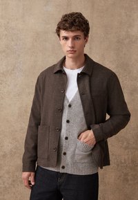 Brown wool jacket with button closure and a chest pocket, layered over a light gray knit cardigan. Model wears dark jeans. Neutral background.