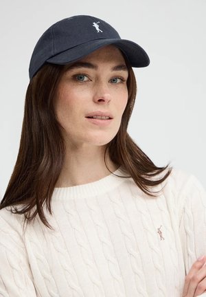 Vrouw met lang bruin haar, een marineblauwe honkbalpet en een witte kabelgebreide trui met een klein geborduurd figuurtje.