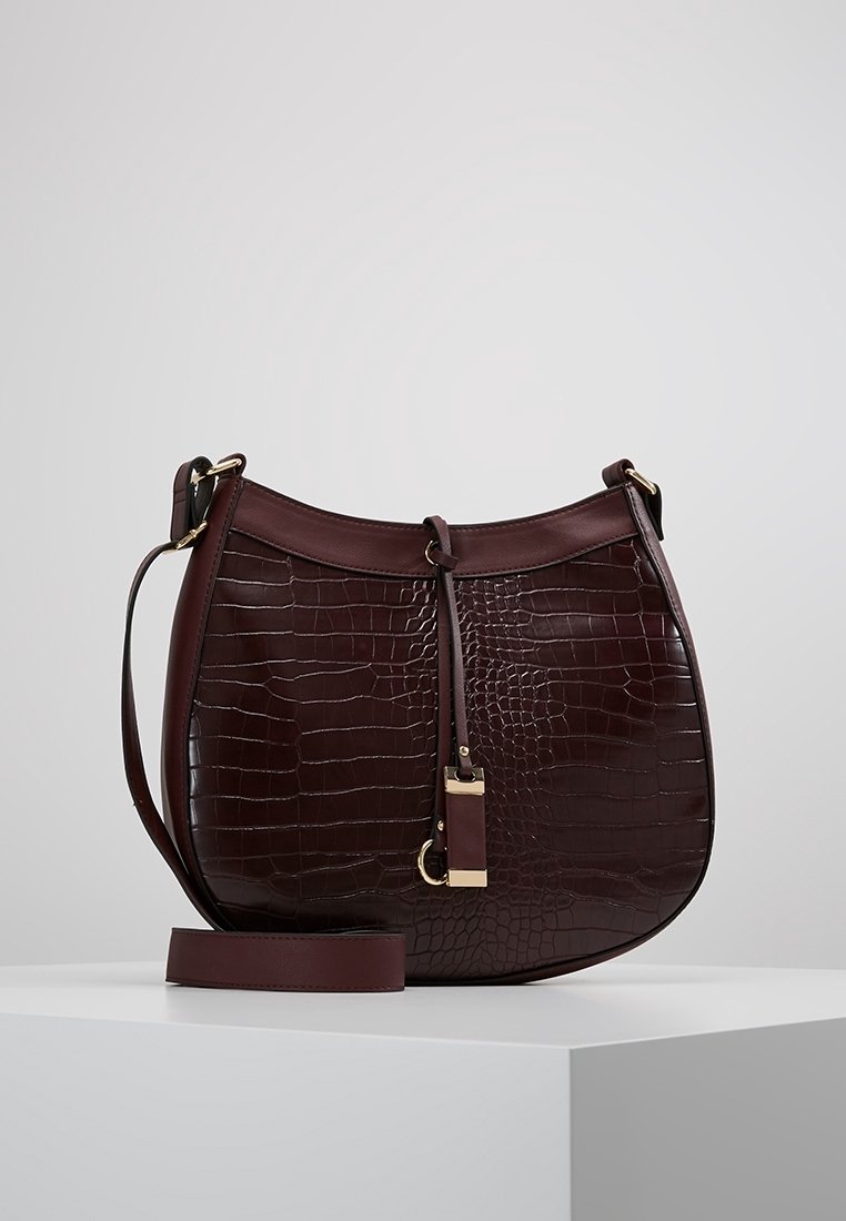 Burgundinpunainen nahkainen käsilaukku krokotiilimallilla, pyöreä muoto, säädettävä olkahihna ja kullanväriset metalliosat.