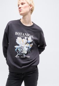 Άτομο που φοράει σκούρο φούτερ με μεγάλο γραφικό μιας μαγκνόλιας σε ανοιχτό μπλε και χρυσό χρώμα και τη λέξη «BOTANICAL» τυπωμένη στο μπροστινό μέρος.