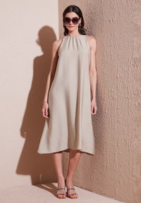 Beige halter dress with a flowing A-line silhouette, textured fabric, and knee-length hem. Paired with strap sandals and oversized sunglasses.