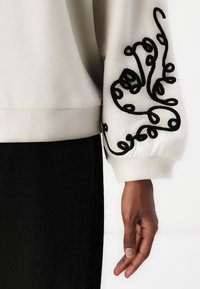 Kremfarget hettegenser med svart svirvlende broderi på ermene, med rund hals og ribbestrikket mansjett på ermet og kant.