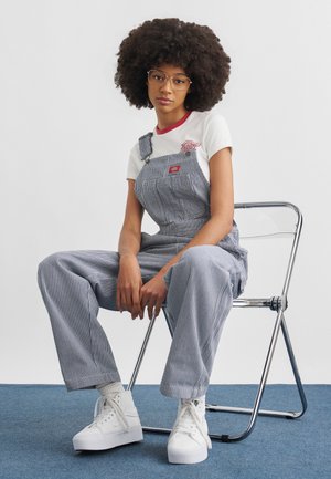 Jonge vrouw met grote afro en bril, gekleed in gestreepte tuinbroek en witte sneakers, zittend op een metalen stoel tegen een witte achtergrond.