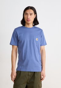 T-shirt in cotone blu con colletto rotondo, maniche corte e una tasca sul petto sinistro con un'etichetta quadrata. Indossato con pantaloni cargo verdi.