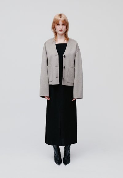 Hellgraue Wollmischungjacke mit boxy Schnitt, übergroßen Ärmeln und drei schwarzen Knöpfen, getragen über einem langen schwarzen Kleid und spitzen schwarzen Stiefeln.