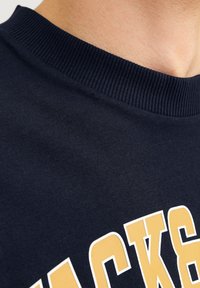 Navy blue cotton sweatshirt featuring a ribbed collar and bold yellow lettering. The fabric has a smooth texture and minimal design details.