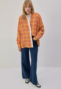 Chemise surdimensionnée à carreaux jaunes et roses avec boutons sur le devant, associée à un jean bleu marine à jambes larges et des baskets vertes. Texture lisse, design décontracté.