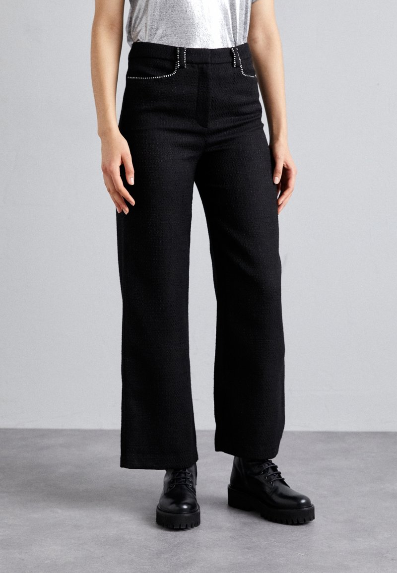 Pantalon noir à jambes larges en tissu texturé, avec une taille haute et des accents métalliques sur les poches, associé à des bottes noires à lacets.
