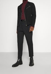 Veste en cuir de moto noire en daim avec des fermetures éclair argentées, col roulé ajusté bordeaux, pantalon noir et bottes Chelsea noires au cuir lisse.