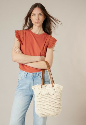 Vrouw met lang bruin haar, gekleed in een oranje mouwloos shirt en lichtblauwe spijkerbroek, met een crèmekleurige gehaakte boodschappentas met leren handvatten in haar hand.