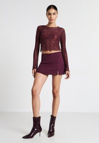 Top corto a maniche lunghe in pizzo bordeaux abbinato a una mini gonna coordinata con una scollatura laterale, completato da stivali alla caviglia lucidi e appuntiti.