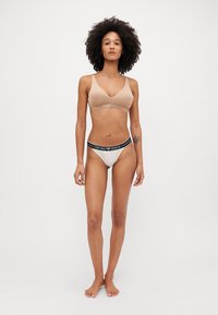 Bralette beige à finition lisse et bretelles ajustables, associé à un string blanc avec une ceinture logo "Emporio Armani" noire.