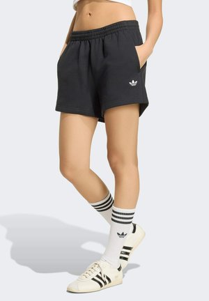 Kvinde iført sorte Adidas-shorts, hvide Adidas-strømper med sorte striber og hvide Adidas-sneakers med sorte striber, stående med hænderne i lommerne.