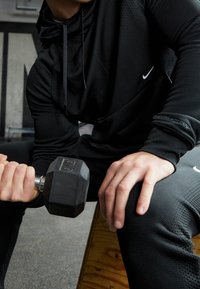 Czarna, teksturowana odzież sportowa z kapturem, trzymająca czarny hantel. Na koszulce widoczny logo Nike oraz dopasowany krój. W tle widoczna drewniana ławka.