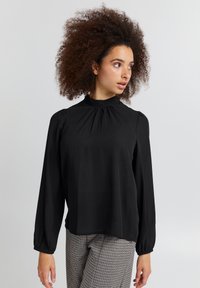Blusa nera a maniche lunghe con collo alto, dettagli arricciati e polsini elastici. Indossata con pantaloni a quadri. Tessuto liscio e leggero.