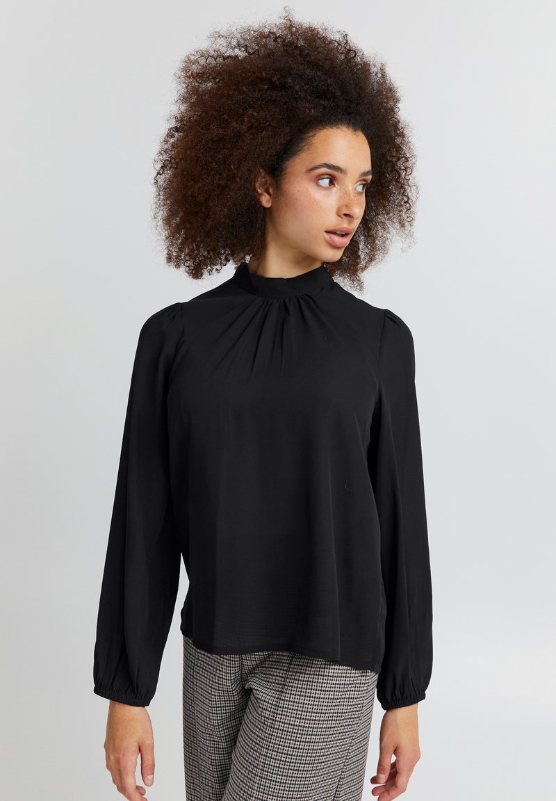 Blusa nera a maniche lunghe con collo alto, dettagli arricciati e polsini elastici. Indossata con pantaloni a quadri. Tessuto liscio e leggero.