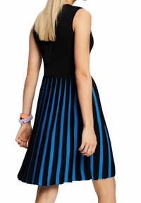 Femme portant une robe noire sans manches avec des rayures plissées verticales bleues et une montre pastel à son poignet gauche.
