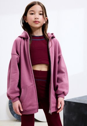 Ung pige iført en lyserød oversized zip-up hættetrøje over en burgundy cropped top og høj-taljede leggings, stående indendørs med en neutral baggrund.