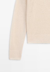 Manche et ourlet de pull en maille couleur crème avec poignets côtelés et bord inférieur, présentés sur un fond blanc.