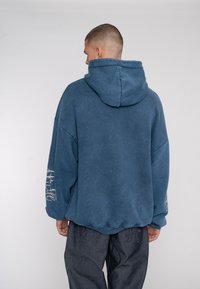 Haut de sweat à capuche bleu surdimensionné avec une finition texturée, doté d'une capuche et de détails brodés sur la manche gauche. Porté avec un pantalon sombre et ample.
