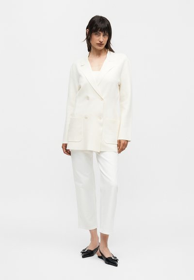 Γυναίκα ντυμένη με λευκό διπλό σακάκι, λευκό κοντό παντελόνι και μαύρα μυτερά φλατ παπούτσια με ασημένιες αγκράφες, στέκεται σε λευκό φόντο.