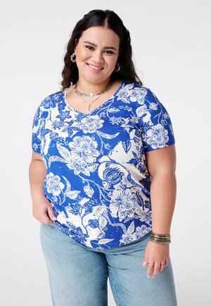 Lachende vrouw met donker haar draagt een blauw-wit bloemenoverhemd, lichtblauwe spijkerbroek, zilveren ketting, creolen en armbanden tegen een witte achtergrond.