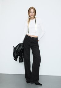 Jeans neri a gamba larga con sottili decorazioni, abbinati a un top aderente bianco a maniche lunghe con dettagli in corda verde e una giacca di pelle nera.