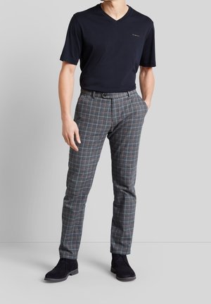 Mężczyzna ubrany w czarną koszulkę z dekoltem w serek, szare spodnie w kratę i czarne eleganckie buty, stoi z jedną ręką w kieszeni na jednolitym tle.