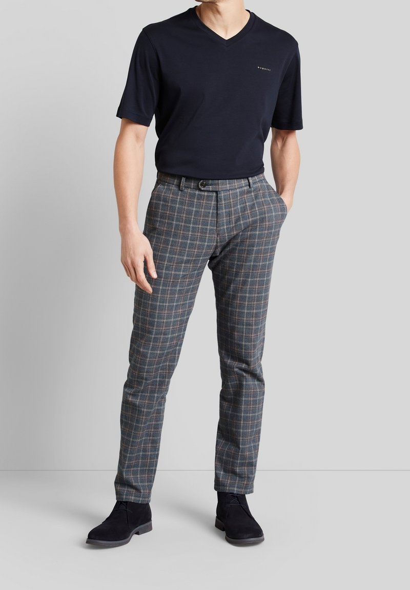 Homme portant un T-shirt noir à col en V, un pantalon écossais gris et des chaussures habillées noires, debout avec une main dans la poche devant un fond uni.