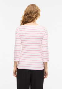 Γυναίκα με κυματιστά ξανθά μαλλιά, που φοράει λευκό μπλουζάκι με μακριά μανίκια και ροζ οριζόντιες ρίγες και μαύρο παντελόνι, με την πλάτη γυρισμένη στην κάμερα.