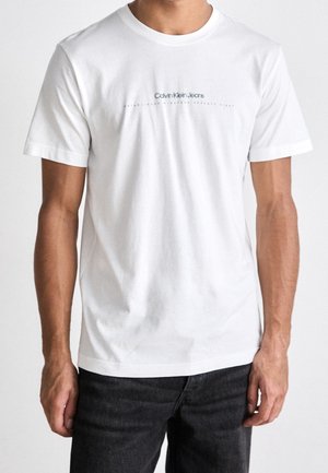Homme portant un t-shirt Calvin Klein Jeans blanc à manches courtes avec un texte noir, assorti à un jean foncé, sur un fond uni.