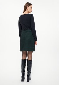 Černý top s dlouhým rukávem v kombinaci se zelenou károvanou sukní. Modelka má na sobě černé vysoké boty do kolen. Hladká tkanina a strukturované designové detaily.