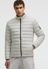 Hellgraue Puffjacke mit horizontalem Steppmuster, hohem Kragen und schwarzem Reißverschluss. Verfügt über enganliegende Bündchen und ein Logo auf der Schulter.