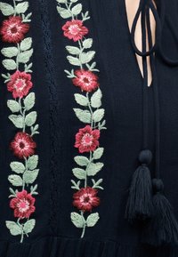 Μαύρο ύφασμα με κάθετη κεντητή διακόσμηση από κόκκινα λουλούδια και πράσινα φύλλα, με λαιμόκοψη με κορδόνια και μαύρα φουντάκια.